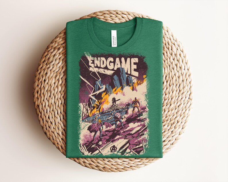 Pu&ograve; includere: Una t-shirt verde bosco con una stampa grafica che riporta il testo ENDGAME in alto. L'illustrazione raffigura una scena di battaglia con diverse figure, terreno roccioso, fulmini e fuoco nelle tonalit&agrave; del viola, arancione e blu.