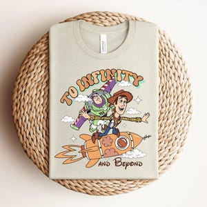 May include: A light beige t-shirt featuring a graphic print of Buzz Lightyear and Woody riding an orange rocket. The text reads TO INFINITY AND BEYOND in brown and orange lettering. The design includes small white clouds and stars on the front.