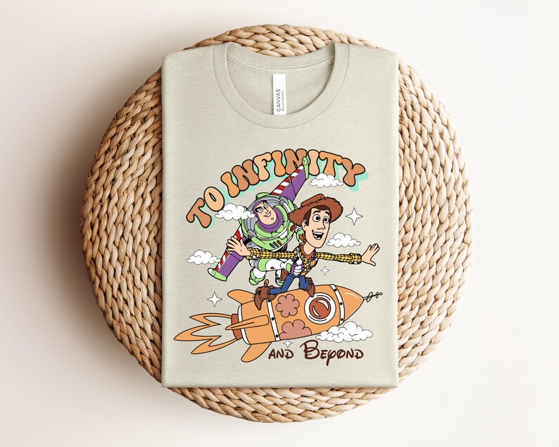 May include: A light beige t-shirt featuring a graphic print of Buzz Lightyear and Woody riding an orange rocket. The text reads TO INFINITY AND BEYOND in brown and orange lettering. The design includes small white clouds and stars on the front.