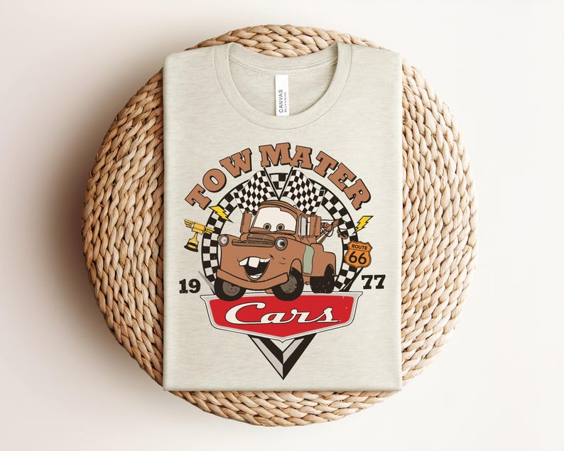 May include: A light beige t-shirt featuring a graphic print of the character Tow Mater from the movie Cars. The design includes a checkered flag, a gold trophy, a Route 66 sign, the text Tow Mater, 1977, and the red Cars logo at the bottom.