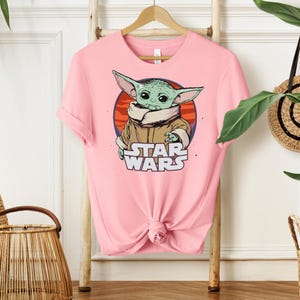 May include: A light pink t-shirt featuring a graphic of a green alien character from Star Wars. The character is set against a red and orange circle, with the words "STAR WARS" below. The shirt is hanging on a wooden hanger.