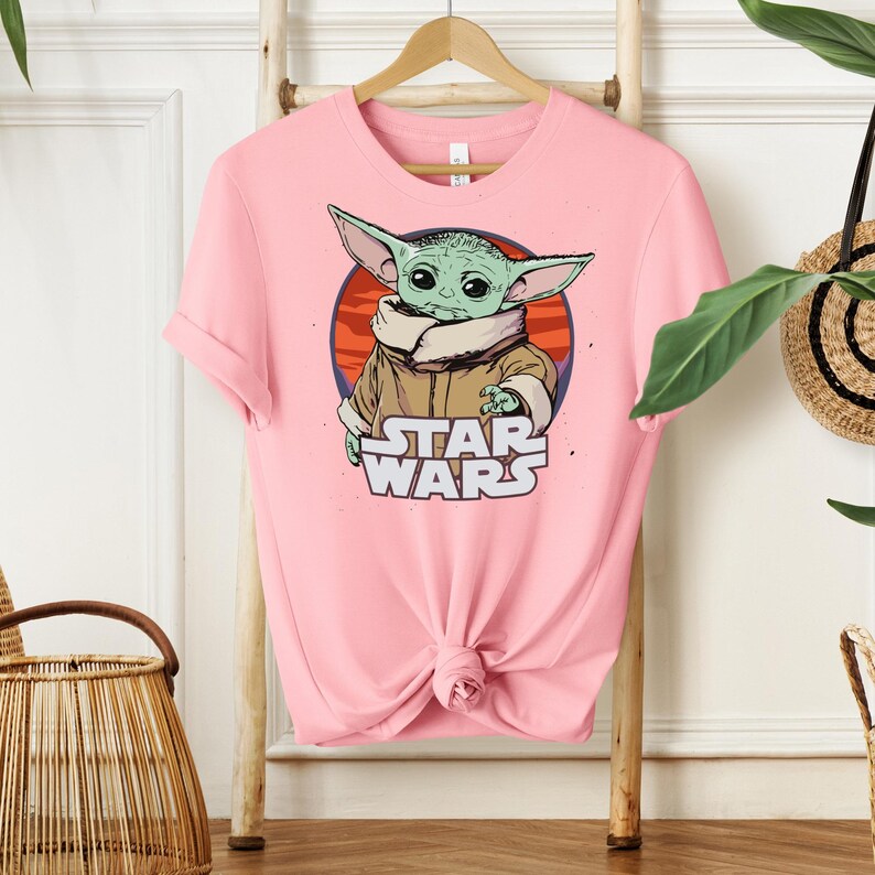 May include: A light pink t-shirt featuring a graphic of a green alien character from Star Wars. The character is set against a red and orange circle, with the words "STAR WARS" below. The shirt is hanging on a wooden hanger.