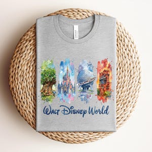 May include: A gray t-shirt featuring watercolor illustrations of iconic Walt Disney World landmarks. The design includes the Tree of Life, Cinderella Castle, Spaceship Earth, and Tower of Terror, with "Walt Disney World" written below.