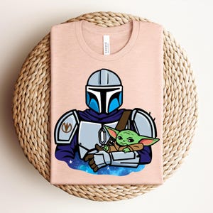Puede incluir: Camiseta rosa claro con una ilustraci&oacute;n de dibujos animados de un personaje mandaloriano sosteniendo a un alien&iacute;gena verde. El mandaloriano lleva un casco y una armadura. El alien&iacute;gena est&aacute; envuelto en una manta marr&oacute;n. La camiseta est&aacute; doblada sobre una superficie tejida.
