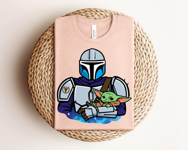 Puede incluir: Camiseta rosa claro con una ilustraci&oacute;n de dibujos animados de un personaje mandaloriano sosteniendo a un alien&iacute;gena verde. El mandaloriano lleva un casco y una armadura. El alien&iacute;gena est&aacute; envuelto en una manta marr&oacute;n. La camiseta est&aacute; doblada sobre una superficie tejida.