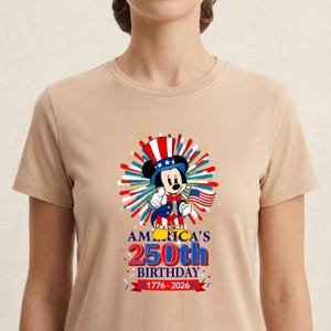 May include: Tan t-shirt featuring a graphic of Mickey Mouse dressed in patriotic attire, holding an American flag. The design celebrates America's 250th birthday, with text reading "AMERICA'S 250th BIRTHDAY 1776-2026". The design includes red, white, and blue elements.