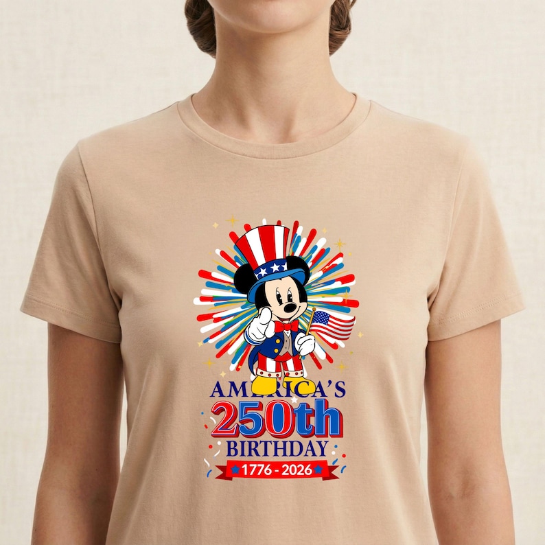 May include: Tan t-shirt featuring a graphic of Mickey Mouse dressed in patriotic attire, holding an American flag. The design celebrates America's 250th birthday, with text reading "AMERICA'S 250th BIRTHDAY 1776-2026". The design includes red, white, and blue elements.