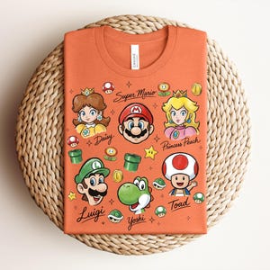 May include: An orange t-shirt featuring cartoon illustrations of Super Mario characters, including Mario, Luigi, Princess Peach, and Yoshi. The text "Super Mario" and character names are also displayed. The shirt is folded on a woven surface.