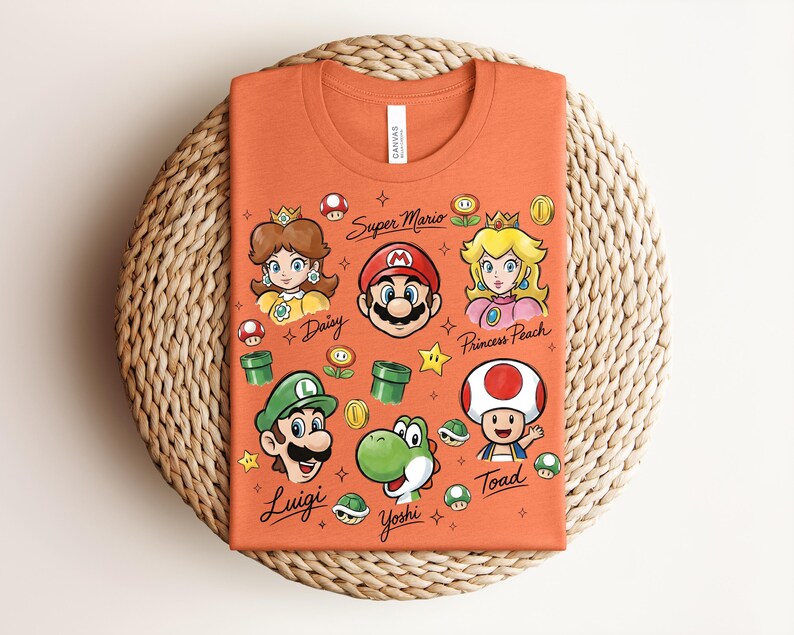 May include: An orange t-shirt featuring cartoon illustrations of Super Mario characters, including Mario, Luigi, Princess Peach, and Yoshi. The text "Super Mario" and character names are also displayed. The shirt is folded on a woven surface.