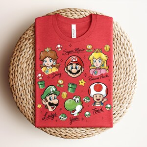 May include: Red t-shirt featuring cartoon characters from Super Mario. The design includes Mario, Princess Peach, Luigi, Yoshi, Toad, and Daisy, along with game-related icons. The words "Super Mario" and character names are also printed on the shirt.
