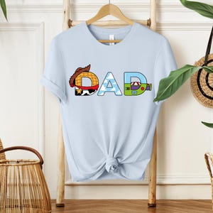 May include: Light blue t-shirt with the word "DAD" in a colorful, cartoon-style design. The letter "D" features a cowboy hat, the "A" has a sky and cloud pattern, and the final "D" has a green and purple design.