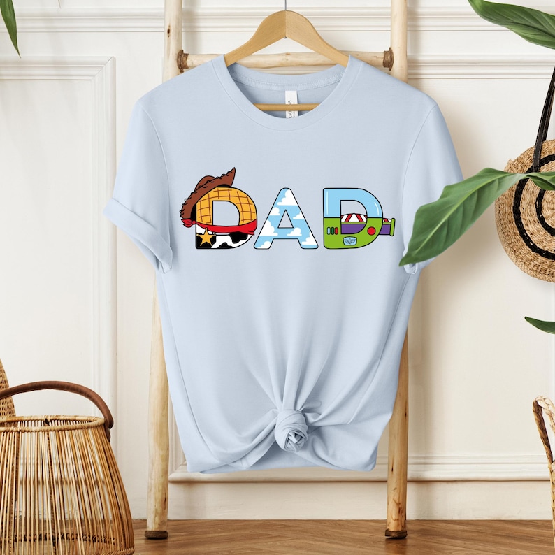 May include: Light blue t-shirt with the word "DAD" in a colorful, cartoon-style design. The letter "D" features a cowboy hat, the "A" has a sky and cloud pattern, and the final "D" has a green and purple design.