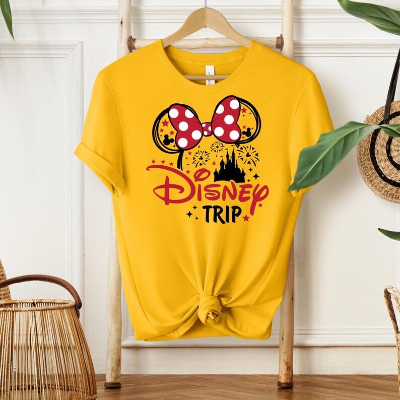 May include: Yellow t-shirt featuring a Minnie Mouse bow with red polka dots and black ears. The shirt has the words "Disney Trip" in red and black, with a castle graphic and fireworks.