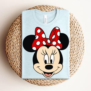 May include: A light blue t-shirt featuring a graphic of Minnie Mouse's face. Minnie's face is black with a red and white polka dot bow. The shirt is folded and placed on a woven, circular surface.