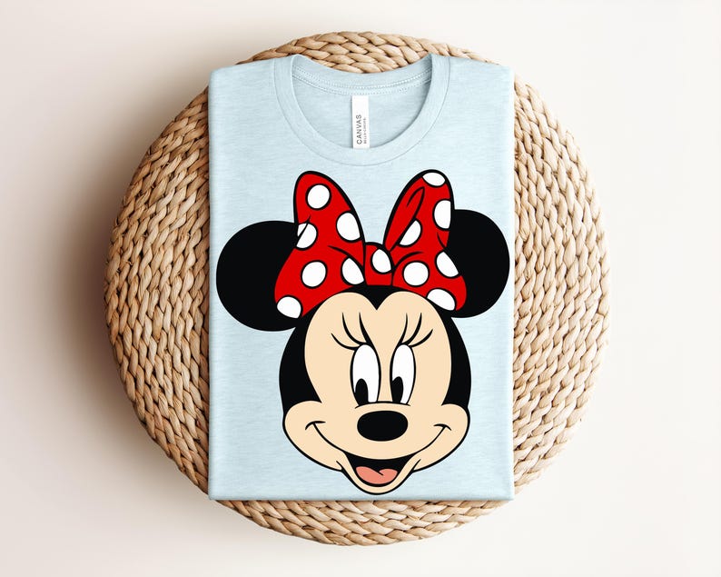 May include: A light blue t-shirt featuring a graphic of Minnie Mouse's face. Minnie's face is black with a red and white polka dot bow. The shirt is folded and placed on a woven, circular surface.