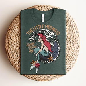 May include: A dark green t-shirt featuring a graphic of a red-haired mermaid with a teal tail sitting on a rock. The design includes white ocean waves, two orange starfish, a pink seashell, and the text THE LITTLE MERMAID Under the Sea '89.