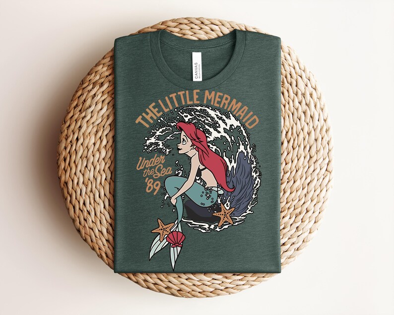 May include: A dark green t-shirt featuring a graphic of a red-haired mermaid with a teal tail sitting on a rock. The design includes white ocean waves, two orange starfish, a pink seashell, and the text THE LITTLE MERMAID Under the Sea '89.