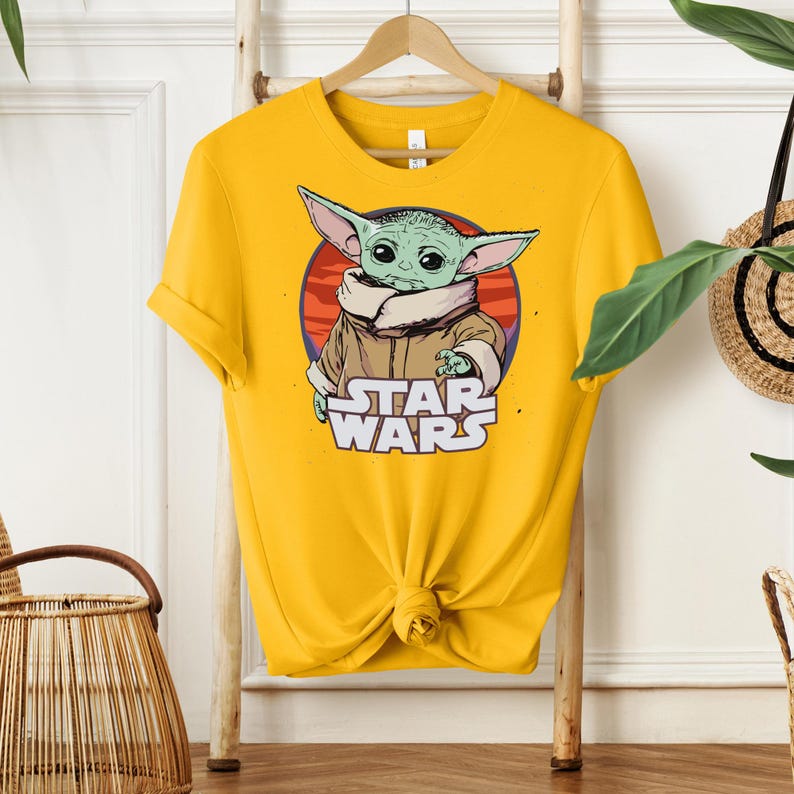 May include: Mustard yellow t-shirt featuring a graphic of Grogu, also known as Baby Yoda, from Star Wars. The design includes Grogu's image in front of a red and orange circle, with the words "STAR WARS" below. The shirt is hanging on a wooden hanger.