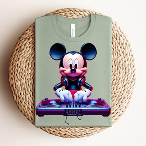 May include: Sage green t-shirt featuring a graphic of Mickey Mouse as a DJ. Mickey is wearing headphones and has his hands on a purple and blue turntable. The shirt is folded on a woven, circular placemat.