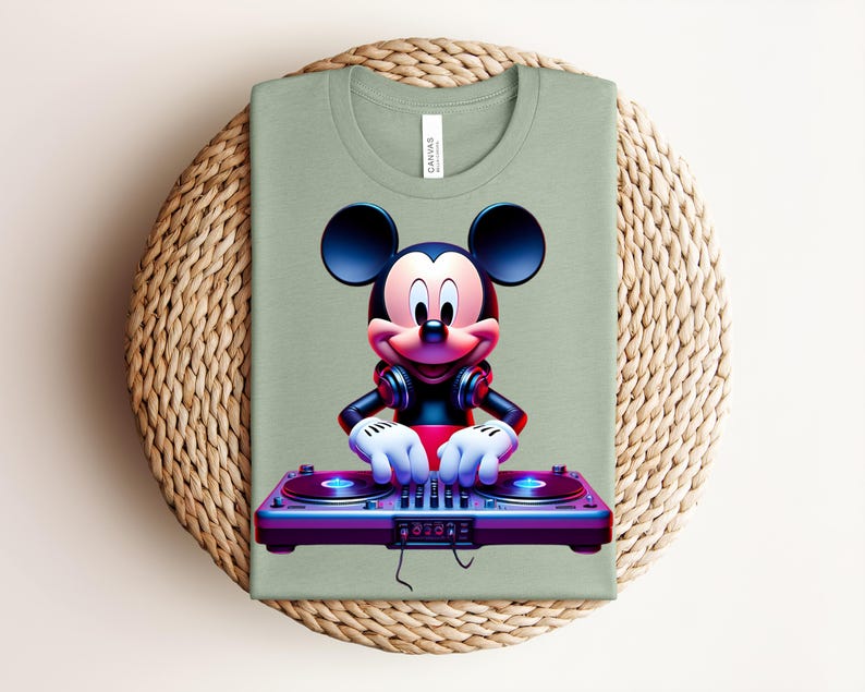 May include: Sage green t-shirt featuring a graphic of Mickey Mouse as a DJ. Mickey is wearing headphones and has his hands on a purple and blue turntable. The shirt is folded on a woven, circular placemat.