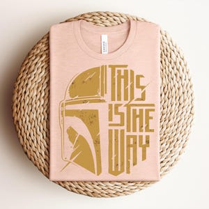 May include: A light peach-colored crew neck t-shirt featuring a distressed gold graphic of a helmet silhouette on the left and the text "THIS IS THE WAY" in a bold, stylized font on the right. The design has a weathered, vintage appearance.