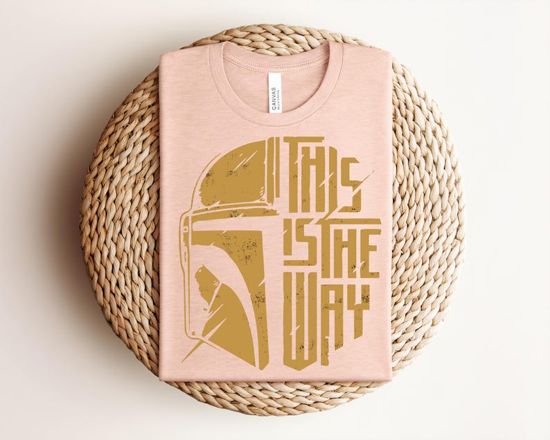 May include: A light peach-colored crew neck t-shirt featuring a distressed gold graphic of a helmet silhouette on the left and the text "THIS IS THE WAY" in a bold, stylized font on the right. The design has a weathered, vintage appearance.