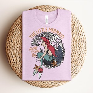 May include: A light purple t-shirt featuring a graphic of a mermaid with long red hair sitting on a rock in front of a large wave. The text reads THE LITTLE MERMAID, Under the Sea, and '89. The design includes two starfish and a seashell.
