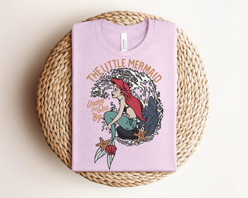 May include: A light purple t-shirt featuring a graphic of a mermaid with long red hair sitting on a rock in front of a large wave. The text reads THE LITTLE MERMAID, Under the Sea, and '89. The design includes two starfish and a seashell.