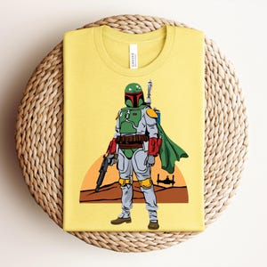 May include: Yellow t-shirt featuring a full-body illustration of a character in green and gray armor, a green cape, and a helmet. The character is holding a black weapon. A sunset scene is in the background. The shirt is folded on a woven surface.
