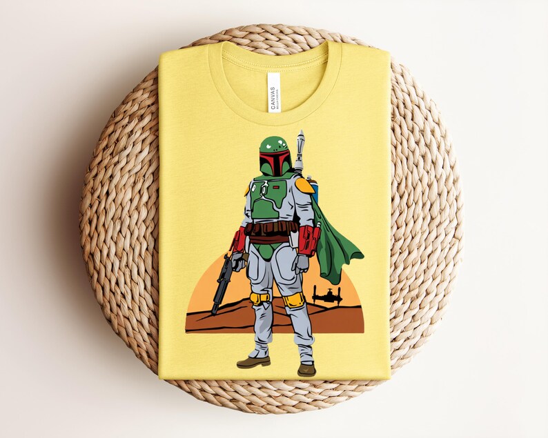 May include: Yellow t-shirt featuring a full-body illustration of a character in green and gray armor, a green cape, and a helmet. The character is holding a black weapon. A sunset scene is in the background. The shirt is folded on a woven surface.