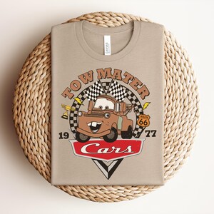 May include: A beige t-shirt featuring a graphic of the character Tow Mater from the movie Cars. The design includes the text Tow Mater, Route 66, and Cars, along with checkered flags, a trophy, and lightning bolts. The numbers 19 and 77 are printed on the sides.