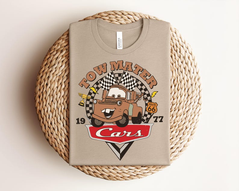 May include: A beige t-shirt featuring a graphic of the character Tow Mater from the movie Cars. The design includes the text Tow Mater, Route 66, and Cars, along with checkered flags, a trophy, and lightning bolts. The numbers 19 and 77 are printed on the sides.