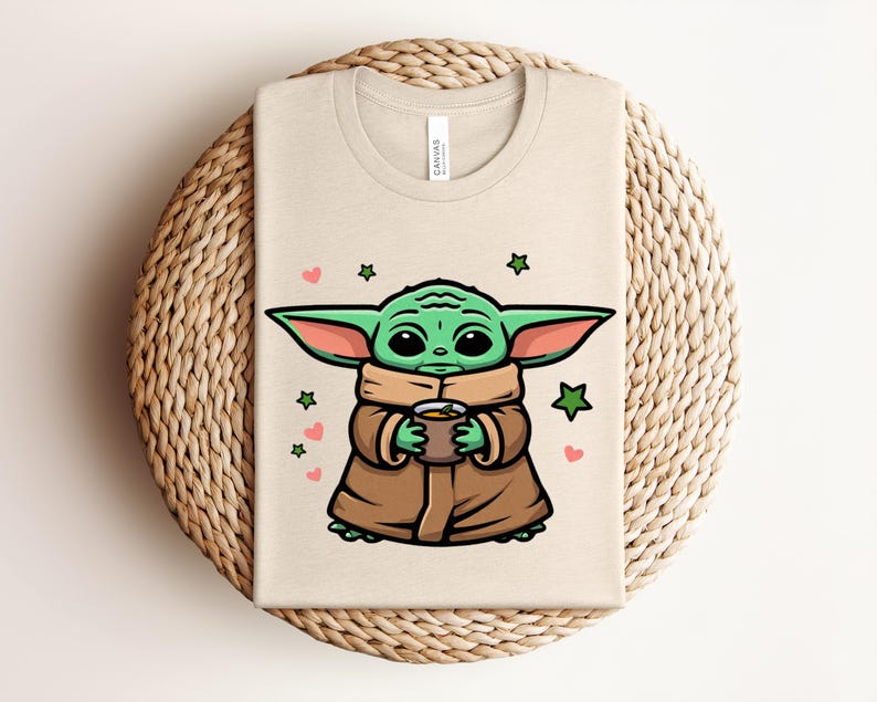 May include: Beige t-shirt featuring a cartoon illustration of a green alien character holding a cup. The design includes green stars and pink hearts. The shirt is folded on a woven, circular surface.