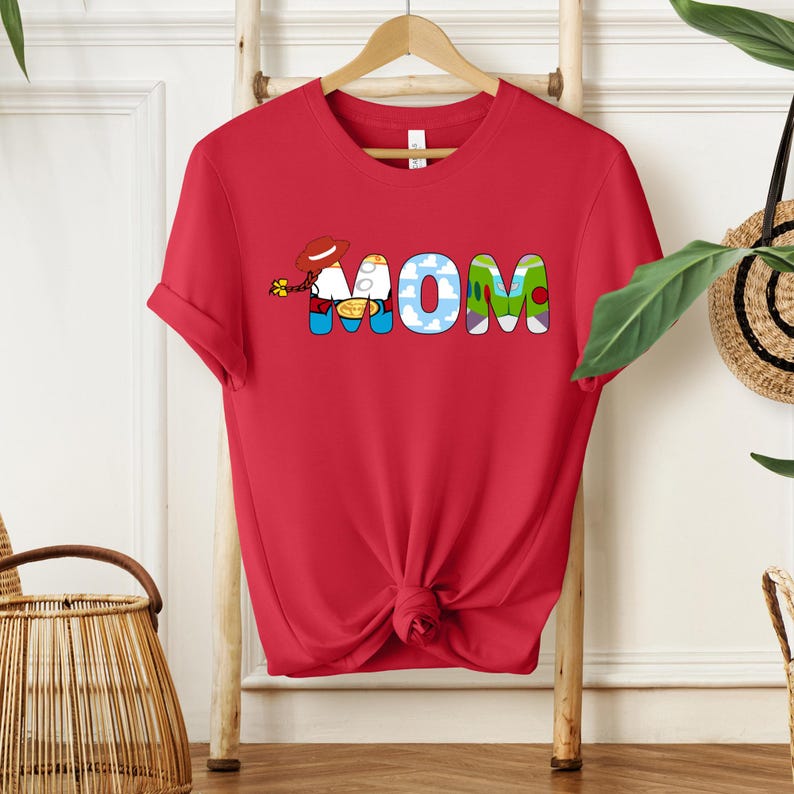 May include: Red t-shirt with the word "MOM" in colorful, cartoon-style letters. The "O" is designed with clouds, and the "M"s have a character with a cowboy hat. A fun and playful design.