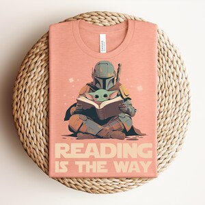 May include: A peach-colored t-shirt featuring a graphic of a helmeted character in armor sitting cross-legged and reading a book with a small green creature. The text below the graphic reads READING IS THE WAY in a bold, sans-serif font.