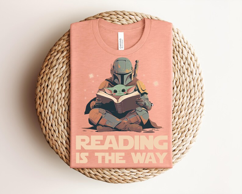May include: A peach-colored t-shirt featuring a graphic of a helmeted character in armor sitting cross-legged and reading a book with a small green creature. The text below the graphic reads READING IS THE WAY in a bold, sans-serif font.