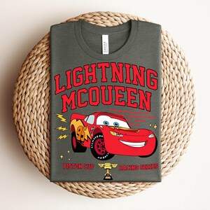 May include: A dark gray t-shirt featuring Lightning McQueen from the Cars movie. The red graphic includes the text "LIGHTNING MCQUEEN" above a red race car with the number 95. Below the car are the words "Piston Cup Racing Series".