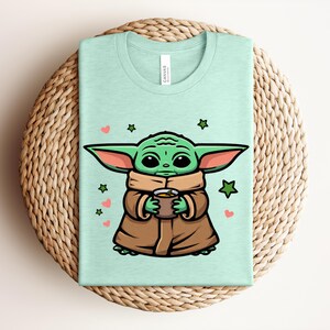 May include: A light green t-shirt featuring a cartoon character with large ears, holding a cup. The character is green with brown clothing, surrounded by hearts and stars. The shirt is folded on a woven surface.