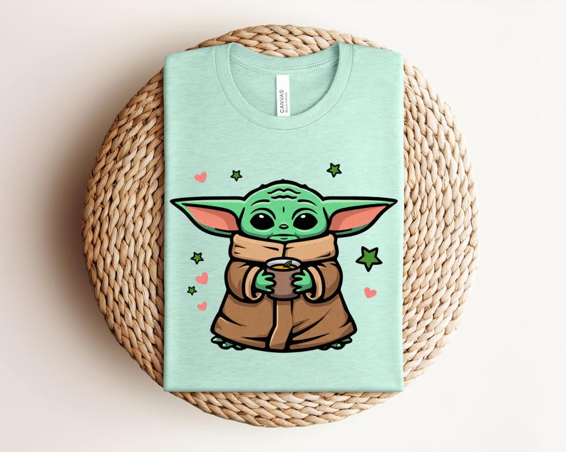 May include: A light green t-shirt featuring a cartoon character with large ears, holding a cup. The character is green with brown clothing, surrounded by hearts and stars. The shirt is folded on a woven surface.