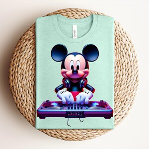 May include: A light green t-shirt featuring a graphic of Mickey Mouse as a DJ. Mickey is wearing headphones and has his hands on a purple DJ mixer. The shirt is folded on a woven, circular surface.