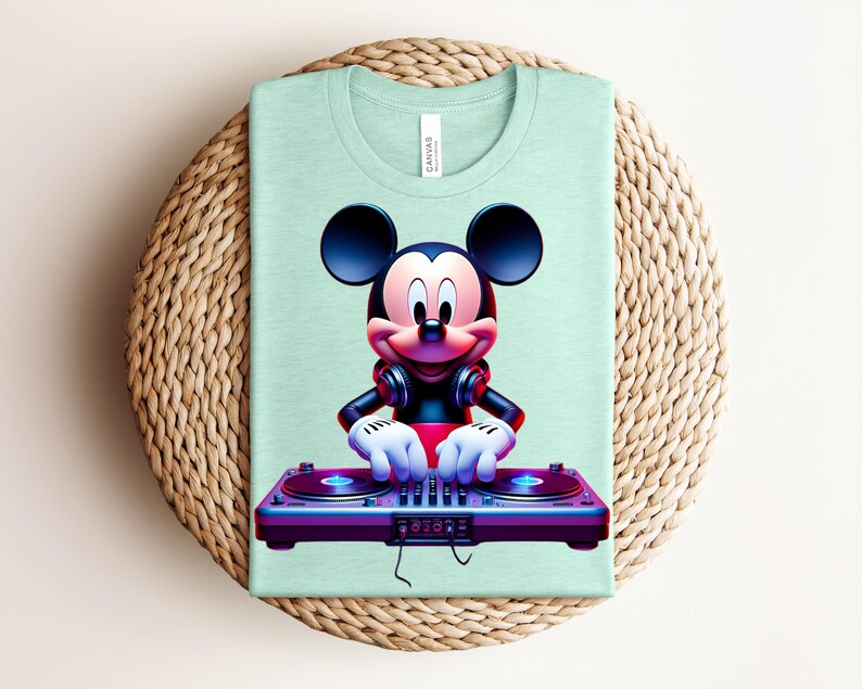 May include: A light green t-shirt featuring a graphic of Mickey Mouse as a DJ. Mickey is wearing headphones and has his hands on a purple DJ mixer. The shirt is folded on a woven, circular surface.