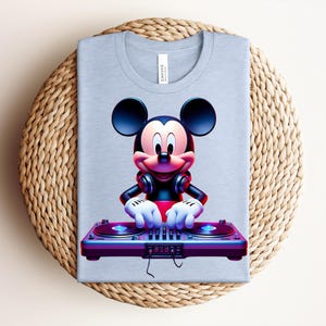 May include: A light blue t-shirt featuring a vibrant graphic of Mickey Mouse as a DJ. Mickey is wearing headphones and operating a turntable. The shirt is folded on a woven, circular surface.