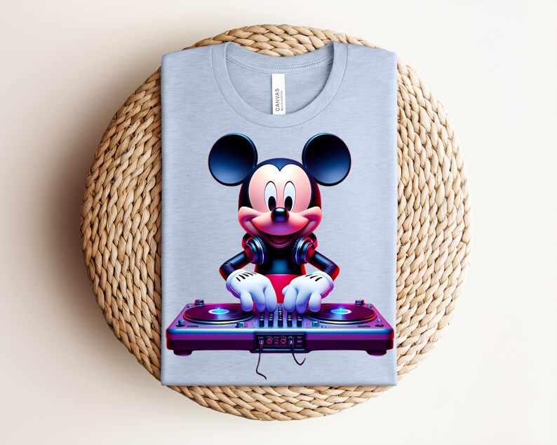 May include: A light blue t-shirt featuring a vibrant graphic of Mickey Mouse as a DJ. Mickey is wearing headphones and operating a turntable. The shirt is folded on a woven, circular surface.
