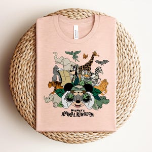Puede incluir: Una camiseta de color melocot&oacute;n claro con un estampado gr&aacute;fico de personajes de Disney con equipo de safari, incluyendo a Mickey Mouse con binoculares, Goofy y varios animales. El texto "DISNEY'S ANIMAL KINGDOM" aparece en negro.