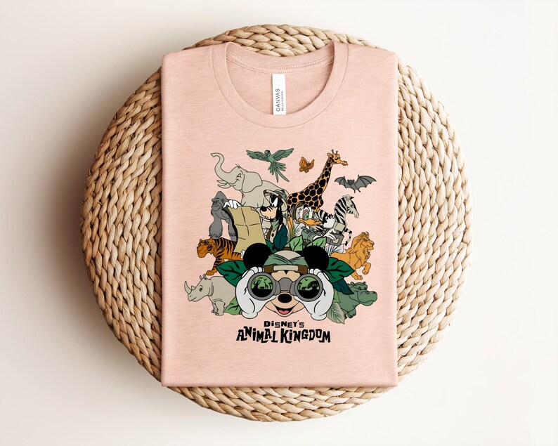 Puede incluir: Una camiseta de color melocot&oacute;n claro con un estampado gr&aacute;fico de personajes de Disney con equipo de safari, incluyendo a Mickey Mouse con binoculares, Goofy y varios animales. El texto "DISNEY'S ANIMAL KINGDOM" aparece en negro.
