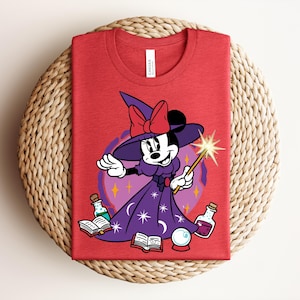 May include: Red t-shirt featuring a cartoon Minnie Mouse dressed as a witch, holding a wand with a starburst. She wears a purple dress and hat with a red bow. The design includes potion bottles, a crystal ball, and open books.