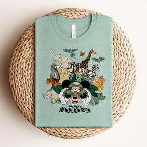 Puede incluir: Una camiseta verde claro con un estampado gr&aacute;fico de Disney's Animal Kingdom. El dise&ntilde;o incluye a Mickey Mouse mirando a trav&eacute;s de binoculares rodeado de varios animales, incluyendo un elefante, jirafa, tigre, cebra y un gorila.