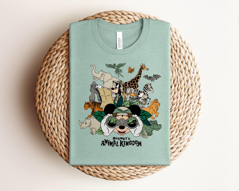 Puede incluir: Una camiseta verde claro con un estampado gr&aacute;fico de Disney's Animal Kingdom. El dise&ntilde;o incluye a Mickey Mouse mirando a trav&eacute;s de binoculares rodeado de varios animales, incluyendo un elefante, jirafa, tigre, cebra y un gorila.