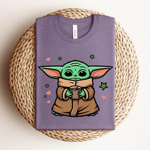 May include: A purple t-shirt featuring a cartoon character holding a cup. The character has green skin, large ears, and is wearing a brown cloak. Pink hearts and green stars are scattered around the character.