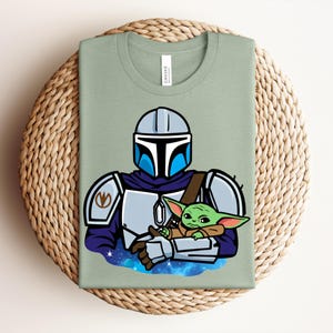 Puede incluir: Camiseta verde salvia con una ilustraci&oacute;n de dibujos animados de un personaje mandaloriano sosteniendo a un alien&iacute;gena verde. El mandaloriano lleva un casco y una armadura plateados con detalles azules. El alien&iacute;gena est&aacute; envuelto en una capa marr&oacute;n.